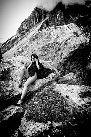 Fashion Glamour photograph of a leggy woman wearing stilettoes, sunglasses and a hooded fur cape, posed on a mountain in the Italian Dolomites, and flashing her coochie.