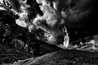 Fine art photo in B&W of a woman in a white flowing gown, posed on knoll agenst a dramatic sky in Wales, UK.