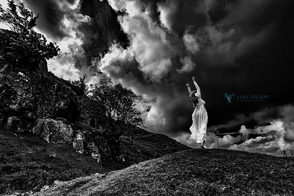 Fine art photo in B&W of a woman in a white flowing gown, posed on knoll agenst a dramatic sky in Wales, UK.