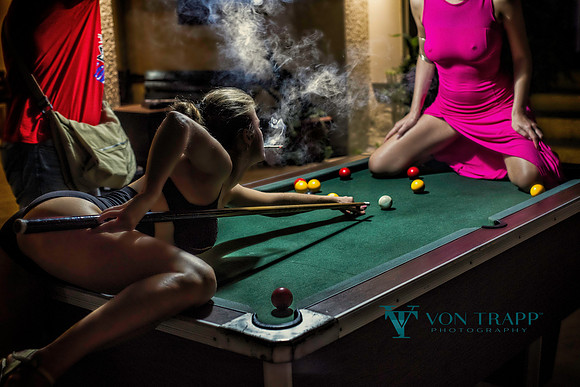 Provocative photo of a woman in a hiked up dress on a pool table while a second woman aims a ball between her legs.