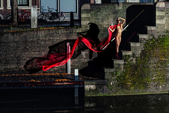 Art nude photo of a woman holding long red fabric blowing in the wind along a canal in Gouda, Holland at night.
