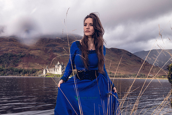 Fine art cosplay photo of a woman in period Gallic dress, posed standing by a lake in front of a Scottish castle.