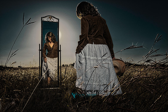 Fine art photo of a lovely woman standing in a field of golden grass, her reflection seen in a full length mirror in the late afternoon.
