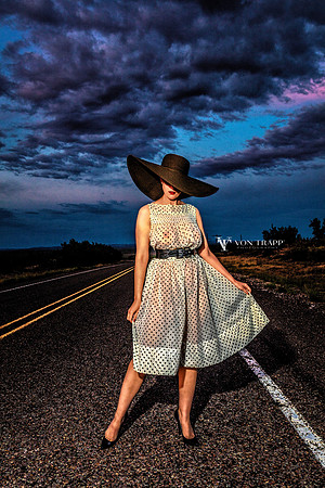 Fashion-Glamour photo of a woman standing on a desert highway wearing a sheer vintage dress, heels and  a broad brim hat.