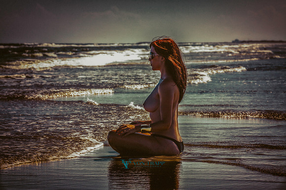 Art nude photo of a topless redheaded woman sitting on a beach at the water's edge. Sexy erotic in public fashion glamour photography.
