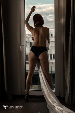 Sexy boudoir photo of a leggy woman wearing panties, holding a white sheet and standing in front of a window.